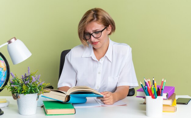 Aluna jovem loira concentrada usando óculos, sentada na mesa com ferramentas escolares, lendo um livro isolado na parede verde oliva