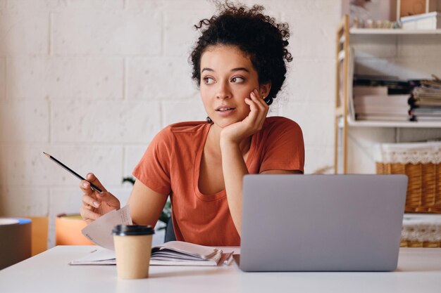 Aluna linda jovem com cabelo escuro encaracolado, sentado à mesa com laptop e xícara de café para ir apoiando-se na mão enquanto sonhadoramente olhando de lado com lápis em casa