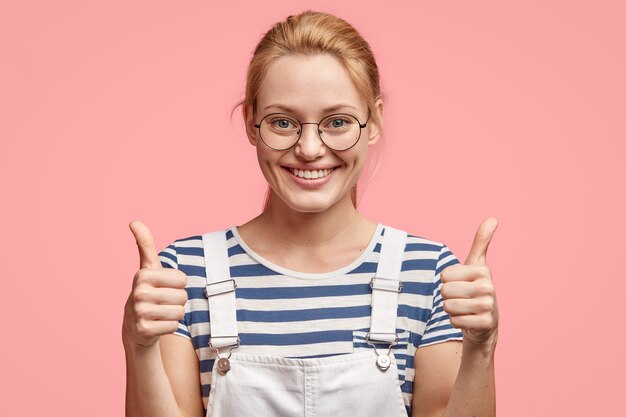 Foto grátis aluna loira alegre e satisfeita desiste de dois polegares, aprova uma nova boa ideia ou conceito de colegas de grupo, tem um sorriso largo