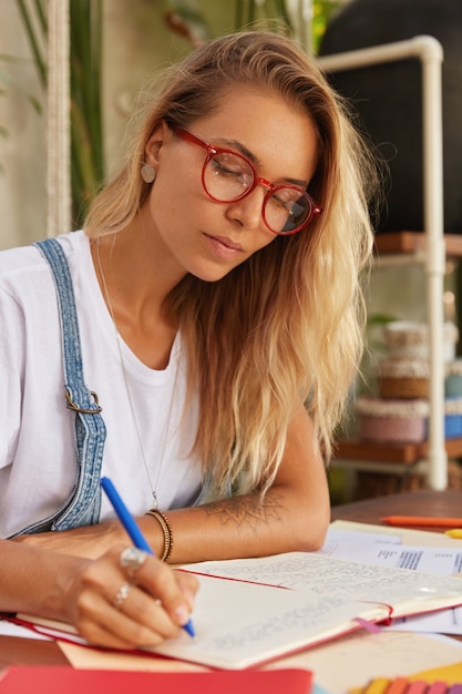 Foto grátis aluna loira com óculos óticos de aro vermelho faz pesquisas para o trabalho do curso