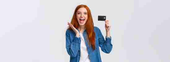 Foto grátis aluna ruiva carismática feliz e bonita em roupas casuais segurando cartão de crédito com viga