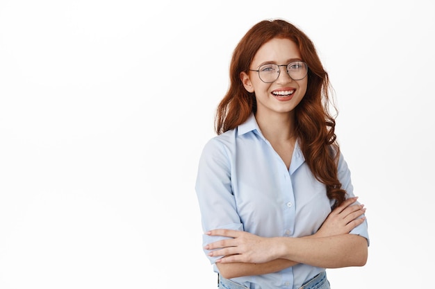 Foto grátis aluna sincera com cabelo ruivo encaracolado, usando óculos e blusa de escritório, cruze os braços no peito, sorrindo feliz e confiante para a câmera, em pé contra um fundo branco