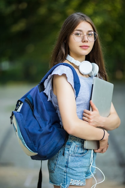 Aluna sorridente com uma mochila segurando um telefone celular, caminhando no parque, ouvindo música com fones de ouvido