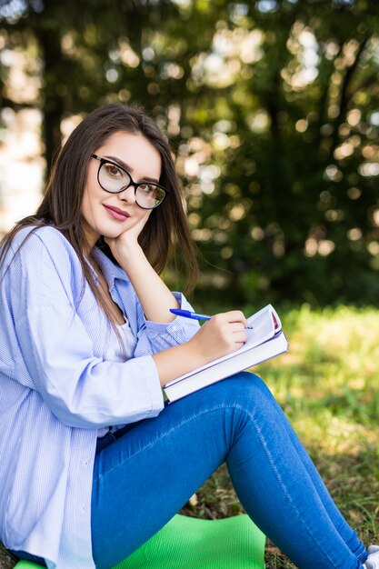 Aluna sorridente de jaqueta jeans e óculos escrevendo no caderno no parque