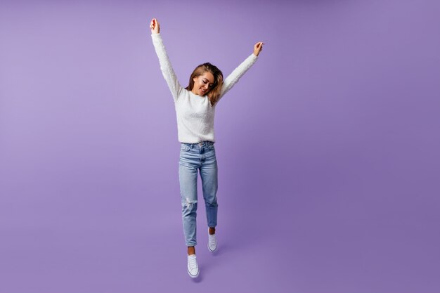 Aluno alegre de bom humor pulando alegremente. Mulher de cabelos longos e cabelos castanhos em jeans elegantes e tênis branco posa para retrato de corpo inteiro