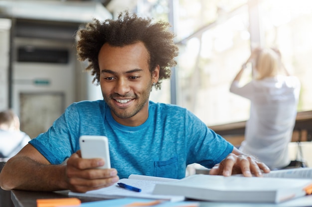 Aluno bonito e alegre usando smartphone, olhando para a tela com um largo sorriso, sentado à mesa do café com um livro didático e um caderno