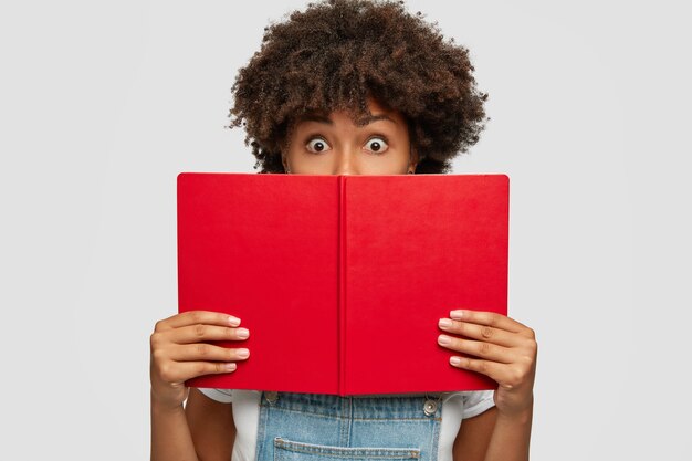 Aluno chocado com corte de cabelo afro, segura livro vermelho aberto na frente, cobre metade do rosto