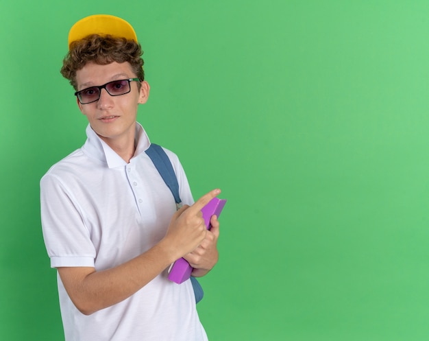 Foto grátis aluno de camisa branca e boné amarelo de óculos com mochila segurando notebook olhando para a câmera sorrindo confiante apontando com o dedo indicador para o espaço da cópia em pé sobre fundo verde