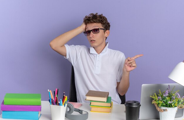 Aluno de camisa pólo branca de óculos, sentado à mesa com livros, parecendo confuso, apontando com o dedo indicador para o lado sobre fundo azul