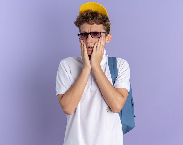 Aluno de camisa pólo branca e boné amarelo usando óculos e mochila olhando para a câmera preocupado em pé sobre um fundo azul