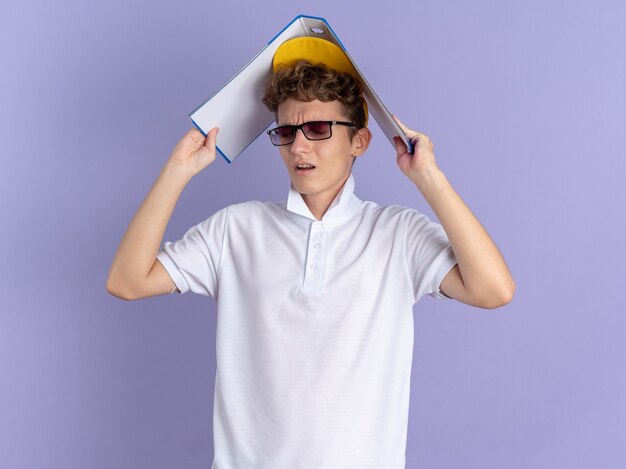 Aluno de camisa pólo branca e boné amarelo, usando óculos, segurando uma pasta