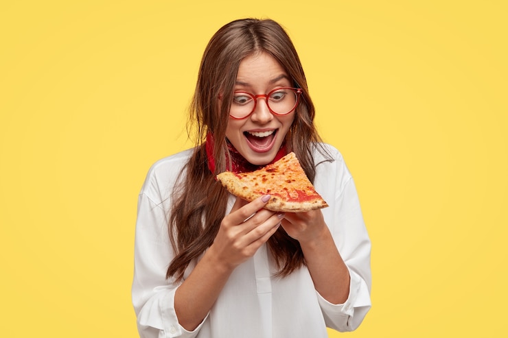 Foto grátis aluno faminto abre amplamente a boca enquanto vê uma deliciosa fatia de pizza, quer comer, vestido de camisa branca, modelos contra a parede amarela. mulher positiva com junk food. pessoas e comendo