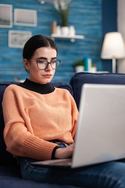 Aluno navegando em informações de comércio on-line usando um laptop enquanto está sentado confortavelmente no sofá da sala de estar. Mulher estudando curso de marketing usando a plataforma da universidade de e-learning