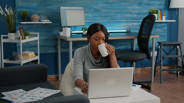 Aluno negro bebendo café digitando artigo nas mídias sociais navegando na webinar de comunicação de palestra no laptop trabalhando na sala de estar