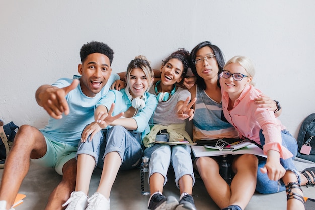 Alunos cansados se preparando para os exames no corredor segurando livros e notas. retrato interno de amigos felizes se abraçando para tirar uma foto enquanto estudam.