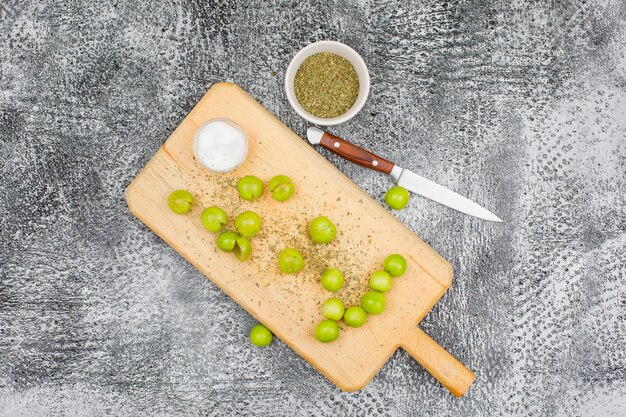 Ameixas verdes picantes em uma tábua com uma pequena barra de sal, tomilho seco e uma vista superior de faca de fruta em uma superfície cinza grunge