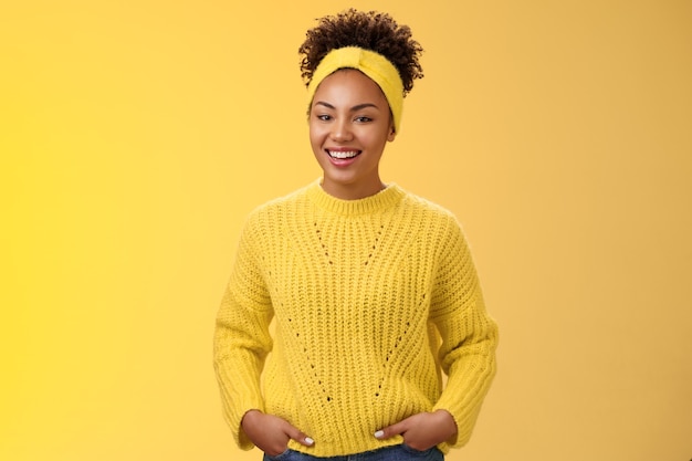 Amigável descontraído jovem afro-americano linda garota headband testa no suéter segurar as mãos nos bolsos sorrindo amplamente se divertir falando uma comunicação interessante, parede amarela de pé.