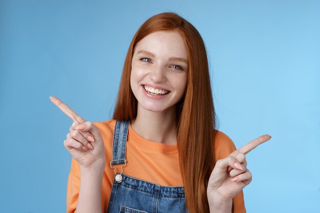 Amigável feliz rindo muito ruiva aluna mostrando muitas oportunidades, conselhos, escolha, apontando para o lado esquerdo para a direita apresentando um produto diferente sorrindo de bom grado, fundo azul.