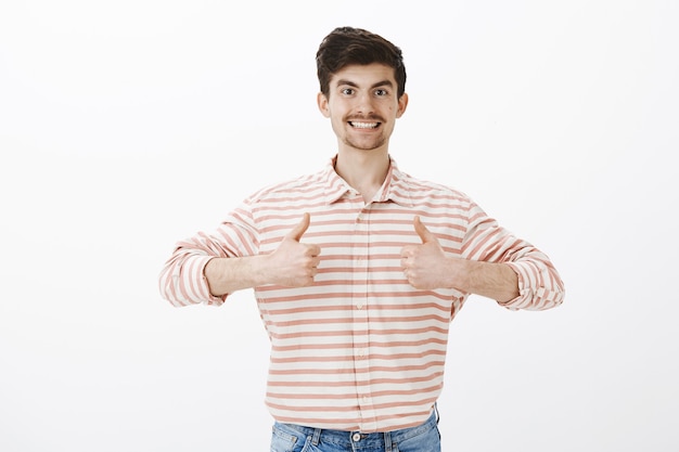 Amigável namorado adulto bonito com camisa listrada, mostrando os polegares para cima e sorrindo feliz, gostando da ideia e dando aprovação, expressando atitude positiva
