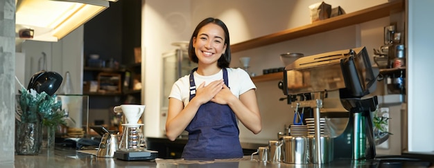 Amigável sorridente garota asiática barista segura as mãos no coração parece grata ao cliente ama seus convidados