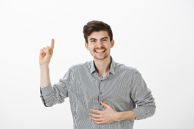 Amigo do sexo masculino positivo e brincalhão com bigode e pintas no rosto, levantando o dedo indicador, segurando a mão na barriga, rindo alto por lembrar o momento engraçado durante a festa sobre a parede cinza