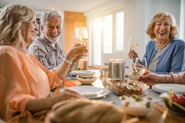 Foto grátis amigos de idosos felizes almoçando juntos e brindando com taças de vinho na mesa de jantar em casa