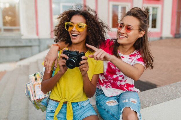 Amigos de meninas felizes sorrindo sentados na rua com uma câmera fotográfica, mulheres se divertindo juntas