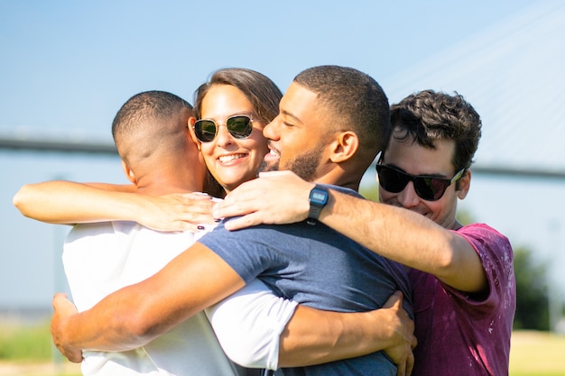 Amigos de sorriso que encontram-se no prado verde durante o dia ensolarado. pessoas alegres, abraçando em círculo no parque. união