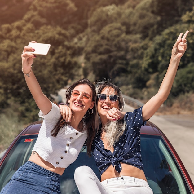 Amigos do sexo feminino felizes sentado no capô do carro, tendo auto-retrato no smartphone