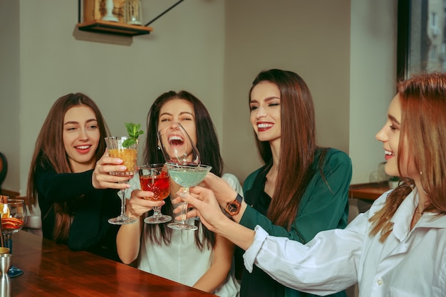 Amigos do sexo feminino tomando uma bebida no bar. Eles estão sentados em uma mesa de madeira com coquetéis. Eles estão batendo os copos