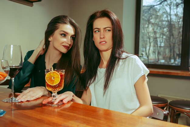 Amigos do sexo feminino tomando uma bebida no bar. Eles estão sentados em uma mesa de madeira com coquetéis. Eles estão vestindo roupas casuais.