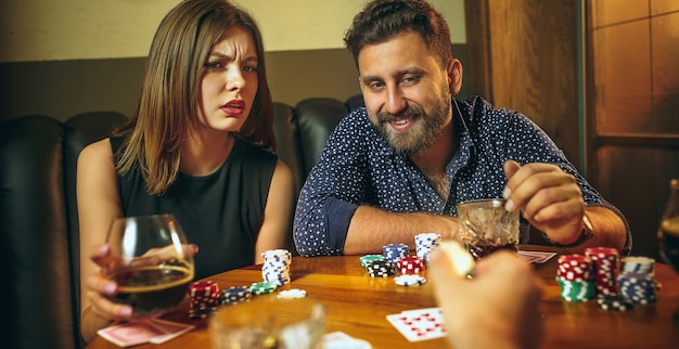 Amigos do sexo masculino e feminino sentados à mesa de madeira. homens e mulheres jogando jogo de cartas. mãos com close-up de álcool. conceito de pôquer, entretenimento noturno e emoção