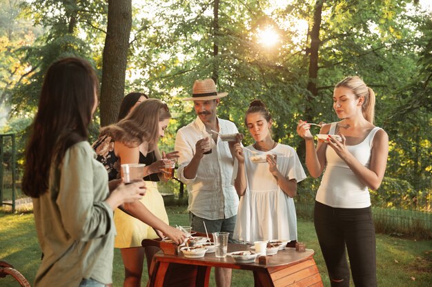 Amigos felizes comendo e bebendo cerveja em um jantar de churrasco na hora do pôr do sol