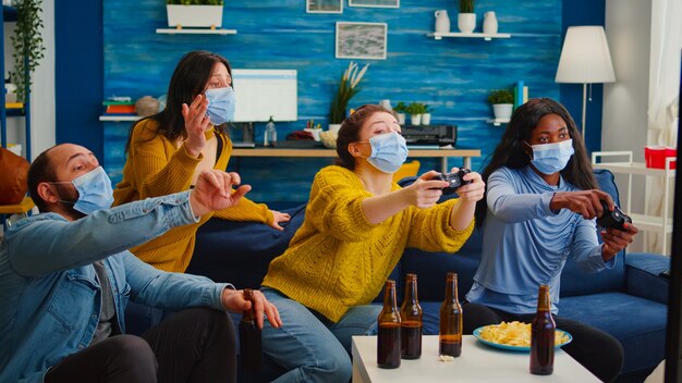 Amigos multiétnicos empolgados tentando ganhar videogames curtindo a nova festa normal durante a pandemia global usando máscara facial, mantendo o distanciamento sentados no sofá na sala de estar apoiando mulheres
