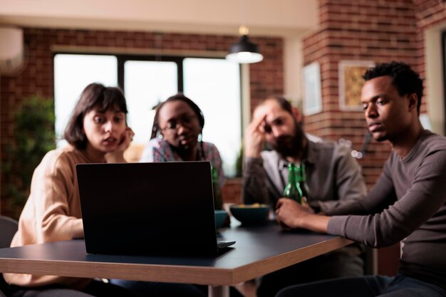 Amigos multiétnicos sentados à mesa enquanto assistem a conteúdo de vídeo no laptop. Grupo diversificado de pessoas em casa na sala de estar curtindo filmes juntos no computador portátil moderno enquanto comem lanches.