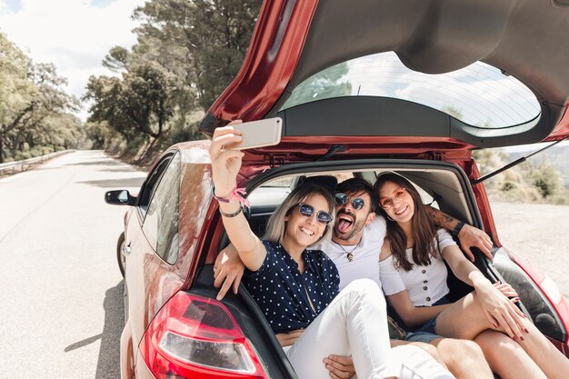 Amigos sentados juntos no porta-malas levando selfie através do telefone móvel