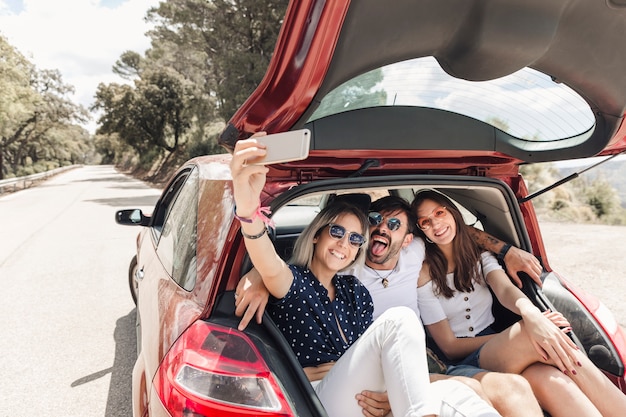 Amigos sentados juntos no porta-malas levando selfie através do telefone móvel