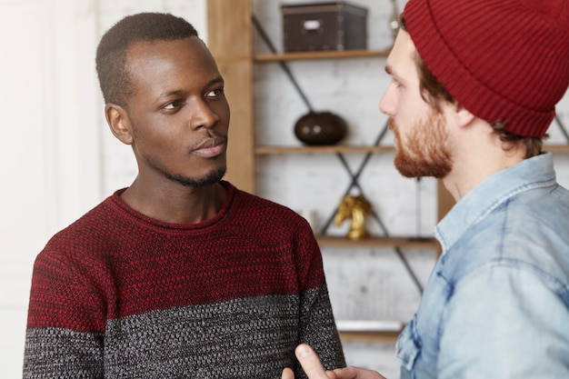 Amizade inter-racial, pessoas, juventude e felicidade. Hipster barbudo elegante de chapéu explicando algo enquanto conversa ou disputa com seu amigo afro-americano vestido de suéter