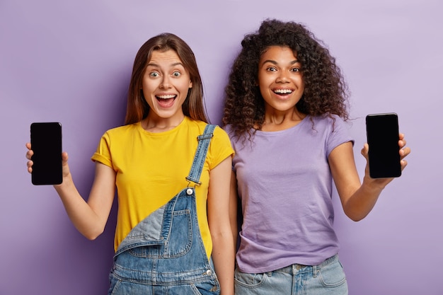 Foto grátis amizade, tecnologia, conceito de publicidade. dois adolescentes multiétnicos sorridentes se aproximam, mostram smartphones com telas de maquete para seu texto