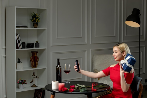 Amor virtual linda garota loira de vestido vermelho na data à distância com vinho segurando presente e telefone