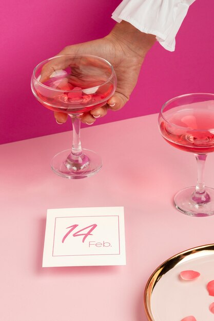 Ângulo alto da mão de uma mulher segurando um copo de bebida ao lado do cartão de dia dos namorados e pétalas de rosa