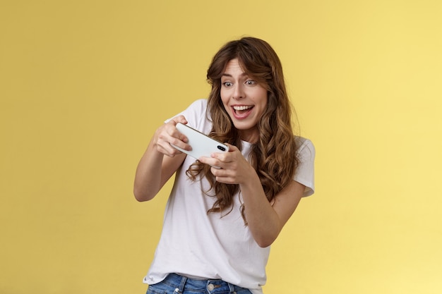 Foto grátis animada, brincalhona, entusiasta, menina, inclinando-se para o lado, jogando incrível jogo de smartphone interessante corrida de carros sorrindo determinado foco de jogo segurar telefone móvel tela de toque horizontal fundo amarelo