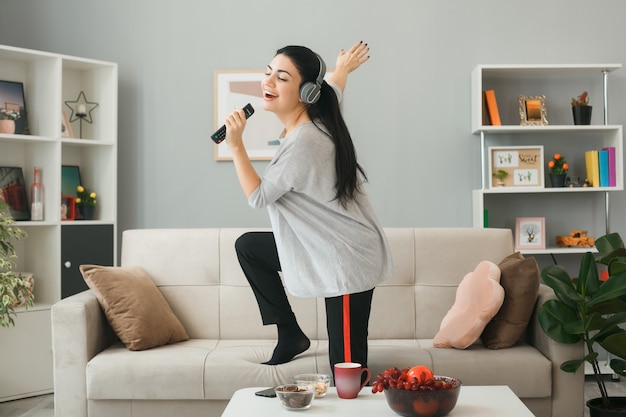 Animada com os olhos fechados, uma jovem usando fones de ouvido segurando o controle remoto da TV canta em pé no sofá atrás da mesa de centro da sala de estar