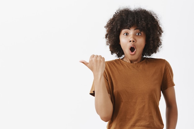 Animada e animada linda garota de pele escura com cabelo encaracolado em uma camiseta marrom da moda apontando para a esquerda com o polegar arfando e dizendo uau com a boca aberta dobrada sendo impressionada fazendo perguntas