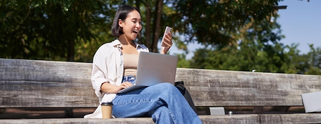 Animada jovem asiática olhando para seu smartphone enquanto está sentada com laptop ao ar livre no parque ensolarado