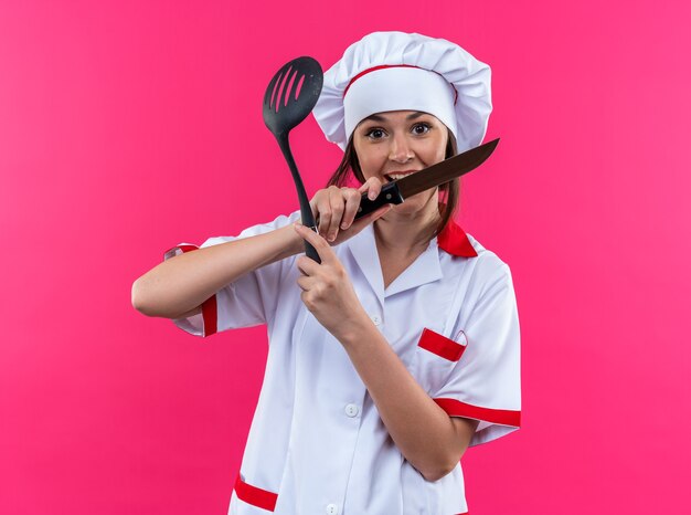 Animada jovem cozinheira vestindo uniforme de chef segurando e cruzando uma faca com uma espátula isolada em um fundo rosa