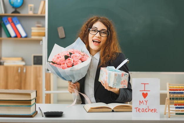 Foto grátis animada jovem professora segurando buquê com presente sentado na mesa com ferramentas escolares em sala de aula