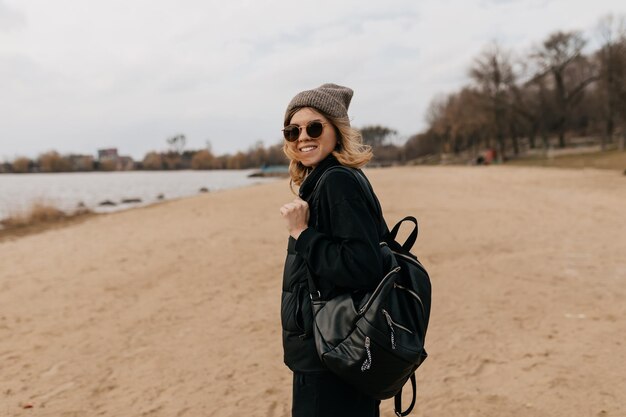 Animada linda garota bonita com penteado curto usando boné com mochila em óculos de sol está andando na praia