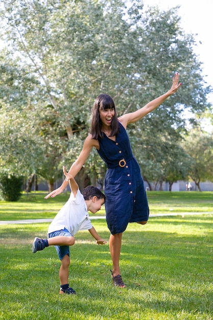 Animada, mãe e filho jogando jogos ativos ao ar livre, em pé e se equilibrando em uma perna, fazendo exercícios engraçados no parque. Conceito de lazer e atividade ao ar livre para a família