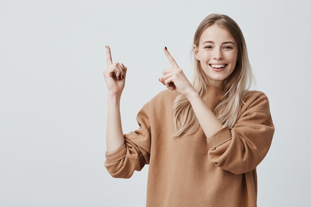 Animado alegre mulher europeia com longos cabelos loiros, vestindo roupas casuais e sorrindo alegremente, apontando os dedos para cima